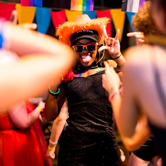 Person wearing an orange hat and sunglasses dances joyfully at a lively party with colorful decorations in the background.