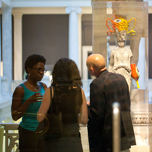Visitors admire a statue adorned with bright colors and unusual headgear, surrounded by elegant museum architecture.