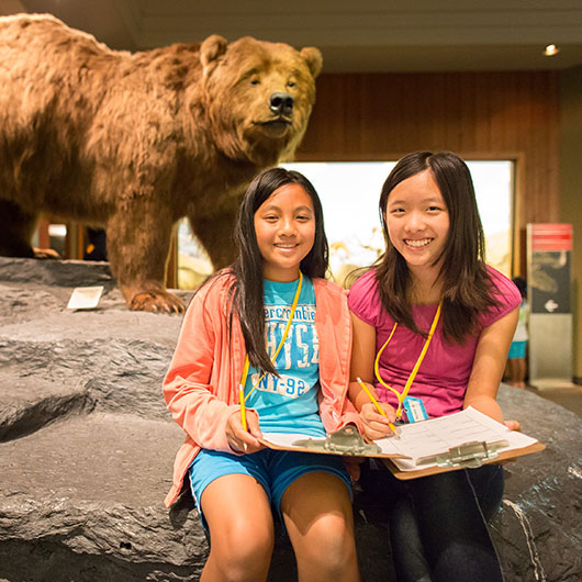 Two children sit on a rock in front of a lifelike bear statue, engaged in drawing and taking notes at an exhibit.