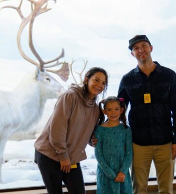 A family poses in front of a reindeer display, set against a snowy background.