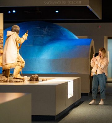 Woman taking a photo of a museum exhibit showing a figure in fur clothing and a blueprint of a snowhouse (igloo) in the background.
