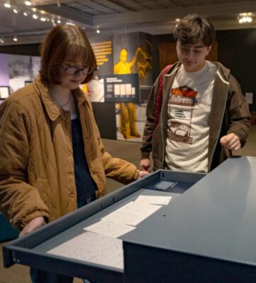 Two individuals examine a display with handwritten notes inside a museum setting, surrounded by informative exhibits.