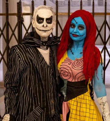A couple dressed in intricate Halloween costumes: one as a skeletal figure in a pinstriped suit, the other as a colorful stitched character.