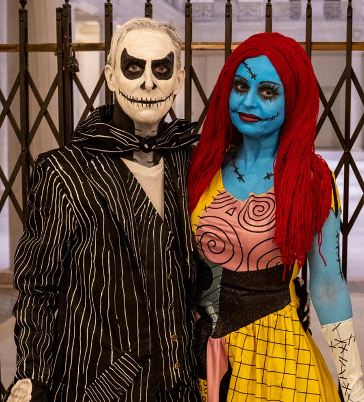 A couple dressed in intricate Halloween costumes: one as a skeletal figure in a pinstriped suit, the other as a colorful stitched character.