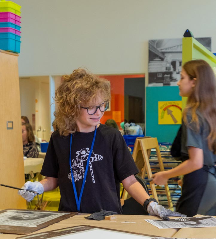 A child with curly hair, wearing a black t-shirt, holds a paintbrush while working on an art project at a vibrant studio.