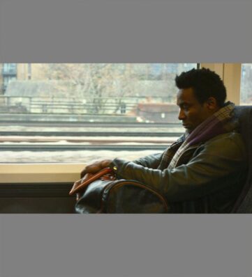 A young black man sitting on a train near a window holding a carry-on.