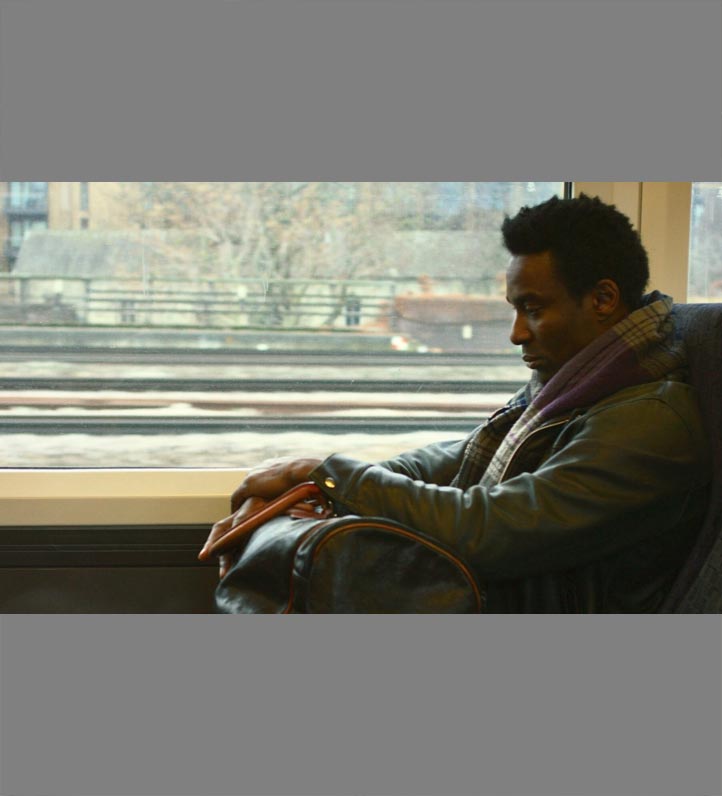 A young black man sitting on a train near a window holding a carry-on.