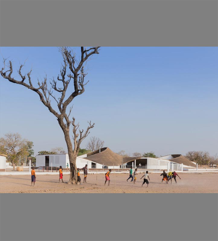 A group of children play in a dusty area near modern white buildings and a tall, bare tree under a clear blue sky.