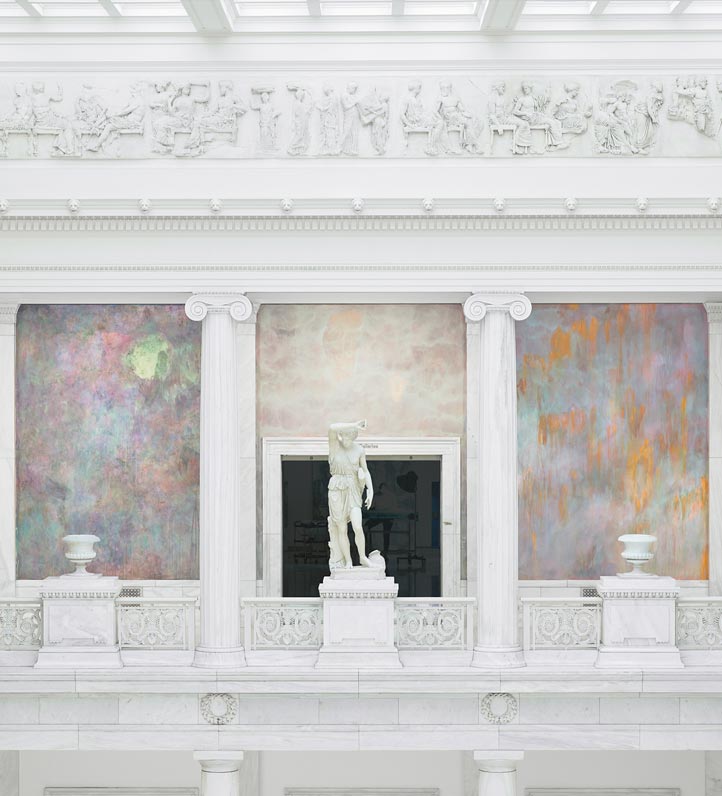 A grand interior featuring marble columns, a white statue at center, and colorful textured walls contrasted against intricate reliefs above.