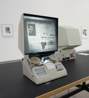 Close-up of a retro news viewing machine displaying old newspaper articles and advertisements, set on a minimalist exhibition table.