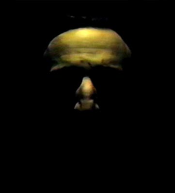 A shadowy face emerges from darkness, illuminated subtly to reveal haunting facial features and a stark expression.