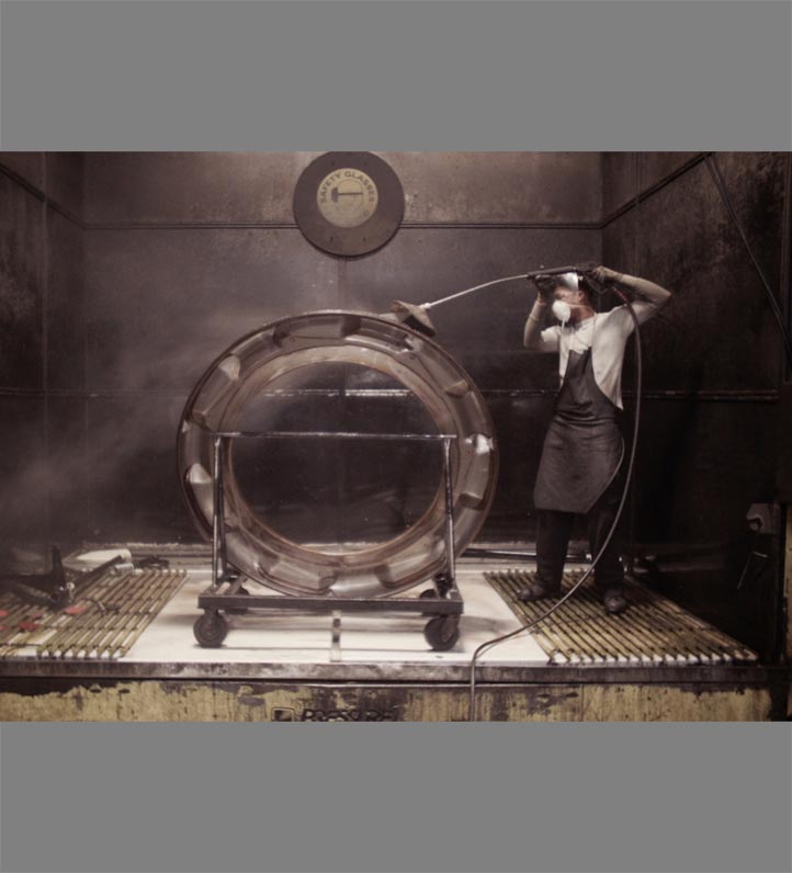 A worker cleans a large circular glass in a misty environment, using a spray tool in an industrial setting.