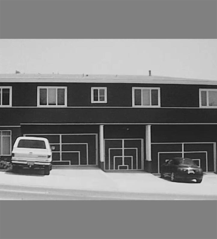 A black and white photo of a two-story building featuring geometric-patterned garage doors, with two parked cars in front.