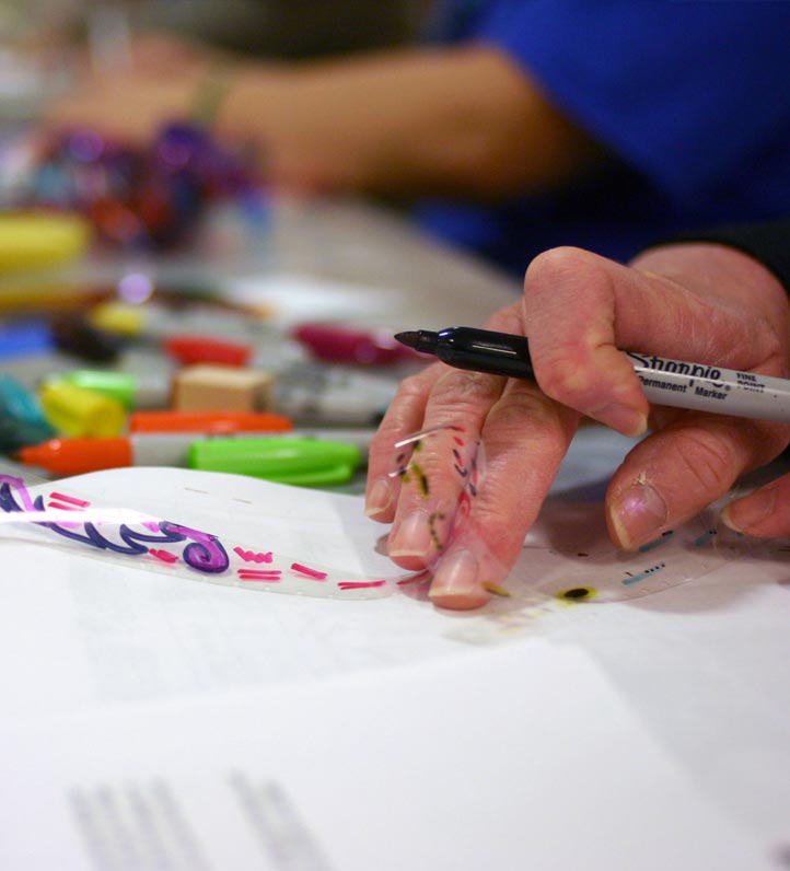 A hand holding a marker decorates clear film with colorful designs, surrounded by various craft supplies on a table.