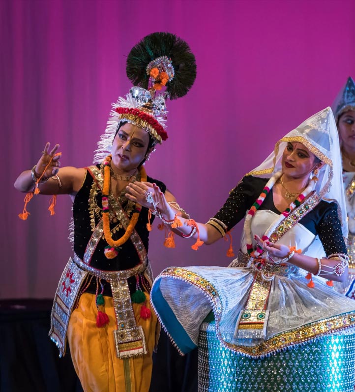 Two dancers in colorful traditional attire perform on stage, showcasing intricate movements against a vibrant purple backdrop.