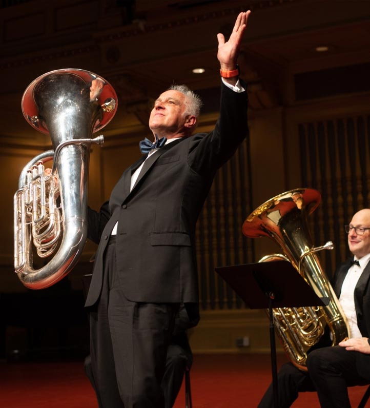 A tuba player in formal wear passionately performs on stage, raising his arm while a second tuba sits on a music stand nearby.