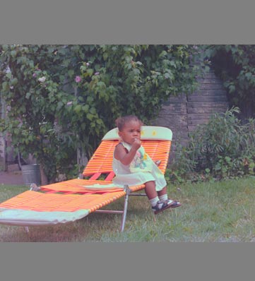 A child relaxes on an orange lounge chair in a lush garden, surrounded by greenery and soft grass.