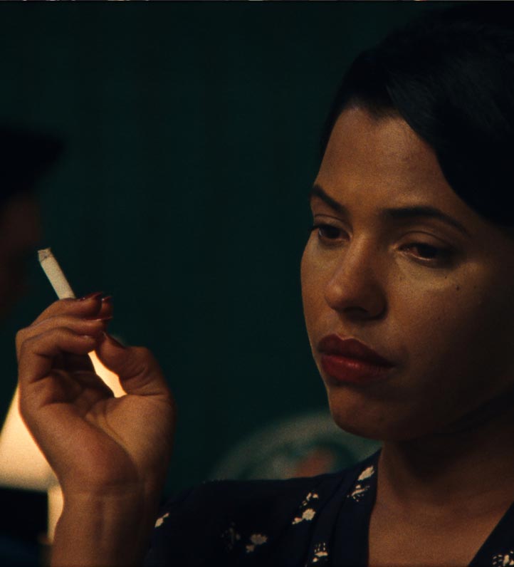 A person holds a cigarette delicately between their fingers, dressed in a patterned garment, with a dark background.