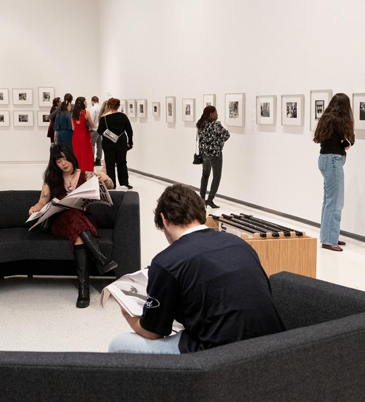 Visitors explore a gallery, viewing framed photographs on display while some engage in reading newspapers while sitting on a modern sofa.