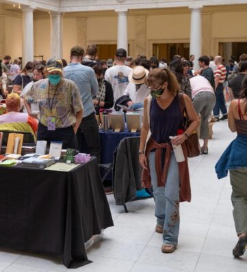 A crowded indoor event filled with people browsing at tables displaying various books, with a woman walking past.