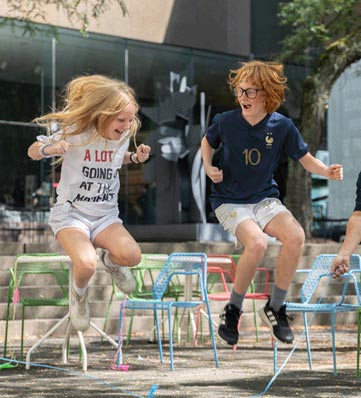Two children jump rope, surrounded by colorful chairs in a sunny outdoor setting.