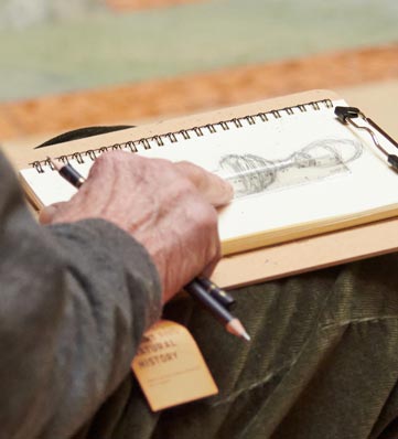 A hand holds a sketchbook featuring a pencil drawing of an hourglass, while seated, with a tag labeled "Visual History" nearby.