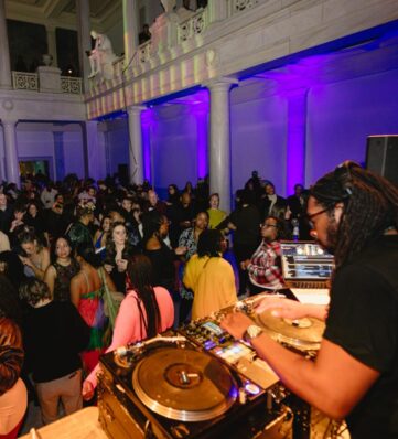 A lively crowd dances in a vibrant venue, illuminated by colorful lights, with a DJ mixing tracks at the foreground.