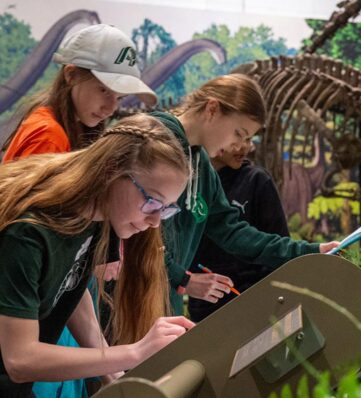 Visitors engage with an interactive exhibit in a dinosaur museum, studying displays amidst lifelike prehistoric scenery.