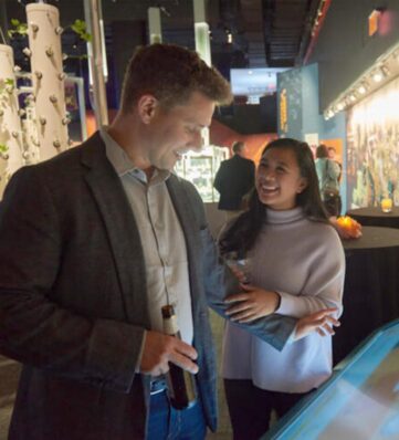 A man and a woman engage with an interactive display at an event, with drinks in hand and a stylish, illuminated background.