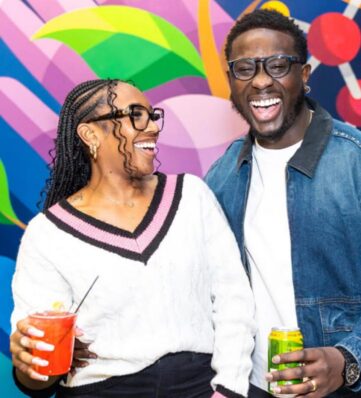 Two people pose together in front of a colorful abstract mural, one holding a brightly colored drink and the other a can.