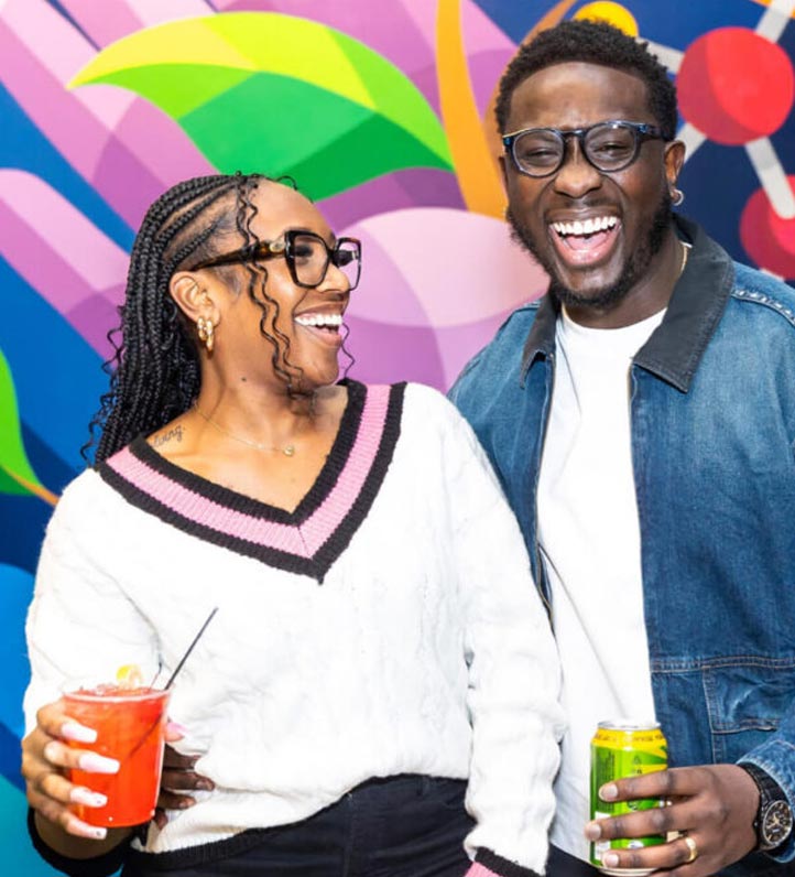 Two people pose together in front of a colorful abstract mural, one holding a brightly colored drink and the other a can.