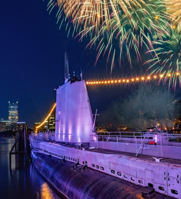 A submarine docked by the river, illuminated by fireworks and city lights, showcasing a festive night scene.