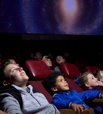 A group of children seated in red theater chairs, captivated by a cosmic projection on a large screen during a planetarium show.