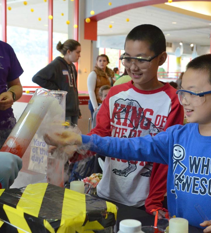 Two children wearing eye goggles holding out their hands to catch a cloud of vapor being poured out of a beaker in a colorful, lively setting.