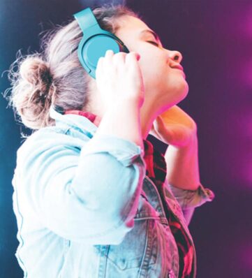 A person enjoying music with teal headphones, wearing a denim jacket surrounded by vibrant purple lighting.