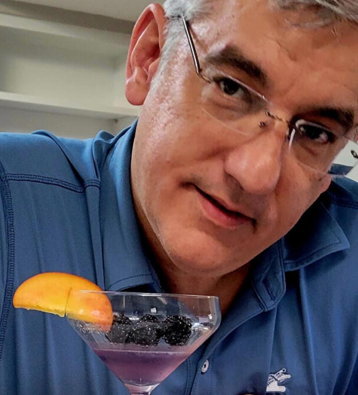 Christopher Monroe, in a blue shirt holds a cocktail glass with a purple drink, garnished with blackberries and an orange slice.
