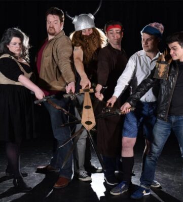A group of six people holding various theatrical swords, dressed in playful costumes, posing dramatically against a dark backdrop.