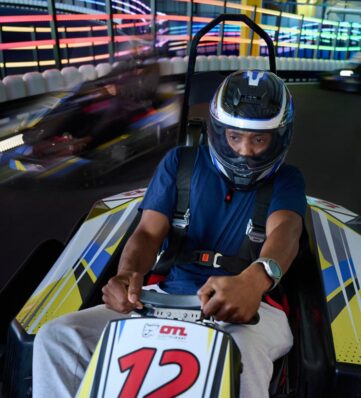 Person wearing helmet driving a go-kart on an indoor neon-lit race track with other racers speeding by.