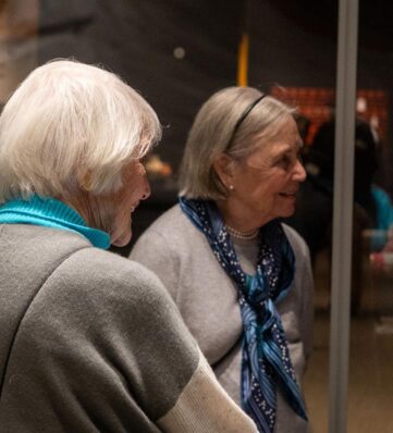 Two older women engage in conversation, one wearing a bright blue scarf and the other a pearl necklace in a warmly lit setting.