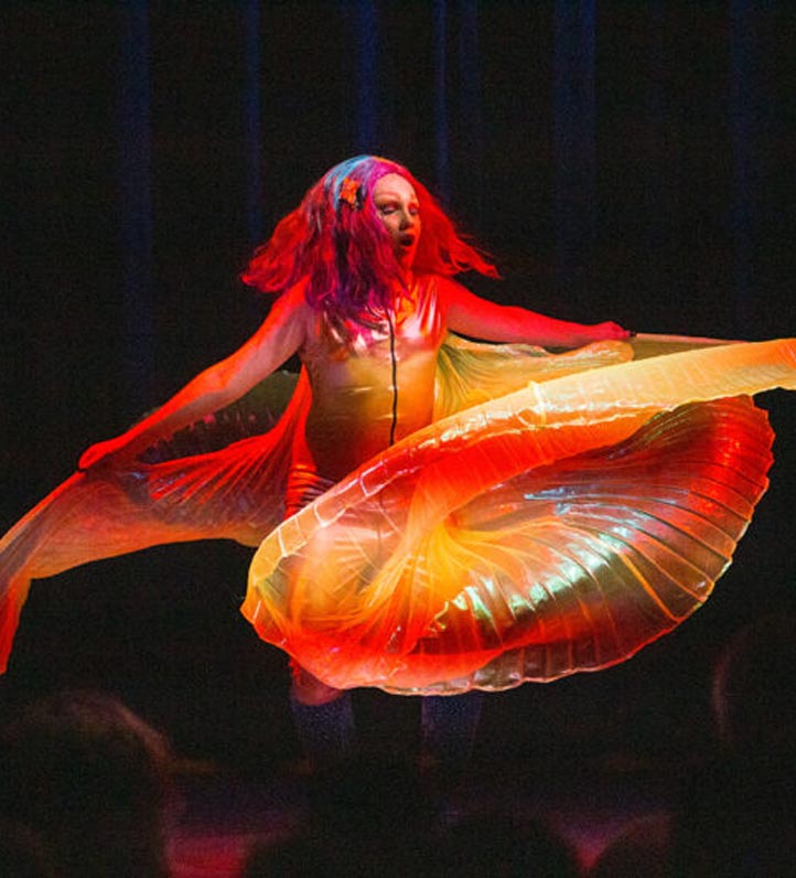 A performer in vibrant, swirling fabric dances under colorful stage lights, showcasing a captivating movement and energy.