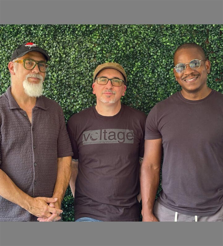 Three men stand together in front of a lush green wall, wearing casual t-shirts and posing confidently for the photo.