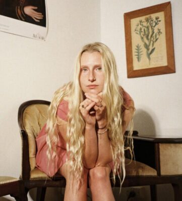 A woman with long, wavy blonde hair sits on a vintage chair, hands clasped, in a warmly lit room adorned with botanical artwork.