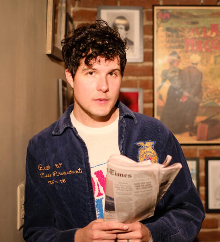 A person stands holding a newspaper in a cozy space adorned with vintage framed pictures and brick walls, wearing a blue corduroy jacket.