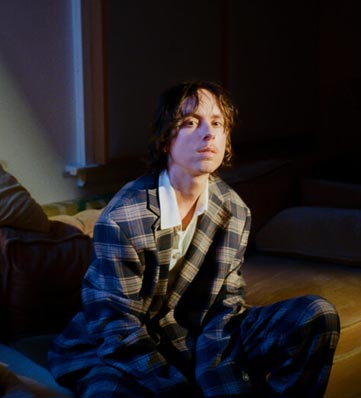A person in a stylish plaid suit sits on a couch, illuminated by soft, warm light, creating an intimate atmosphere.