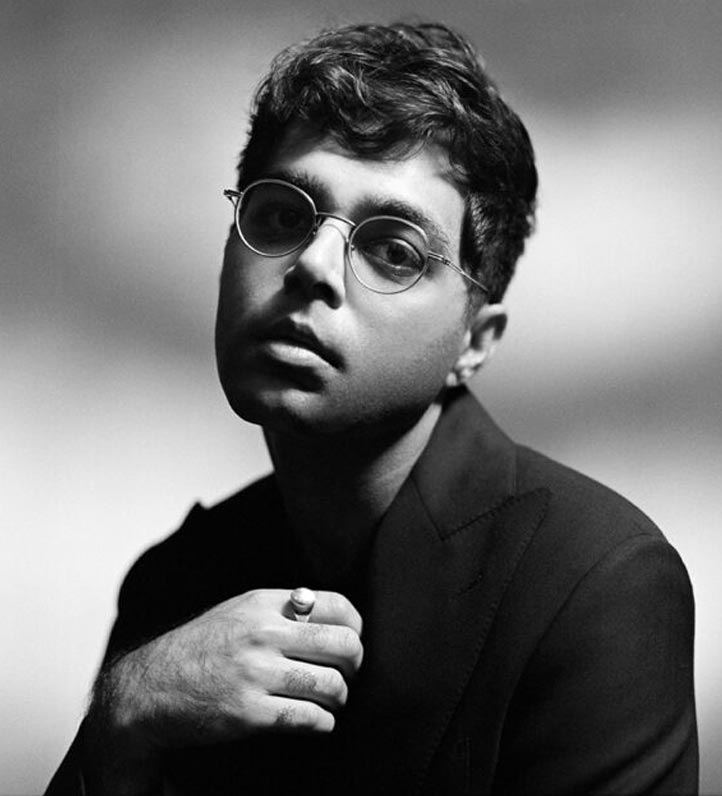 A stylishly dressed man sits in a dramatic black and white setting, gazing thoughtfully.