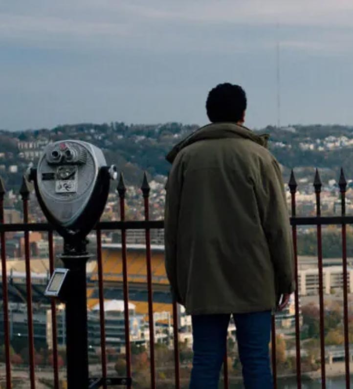 A person stands atop a lookout point, gazing at the Pittsburgh skyline, next to a coin-operated viewing telescope.