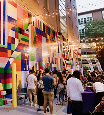 A vibrant outdoor event with a colorful mural backdrop, featuring groups of people socializing and enjoying the atmosphere.