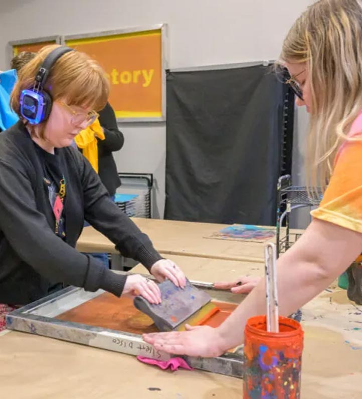 Two people are engaged in screen printing, with vibrant paint and tools on a workspace. One uses a squeegee while the other assists.