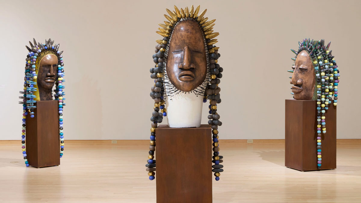 Three large wooden sculpted faces with colorful beaded and pointed headdresses, displayed on pedestals in a gallery room.