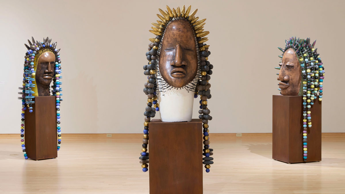 Three large wooden sculpted faces with colorful beaded and pointed headdresses, displayed on pedestals in a gallery room.
